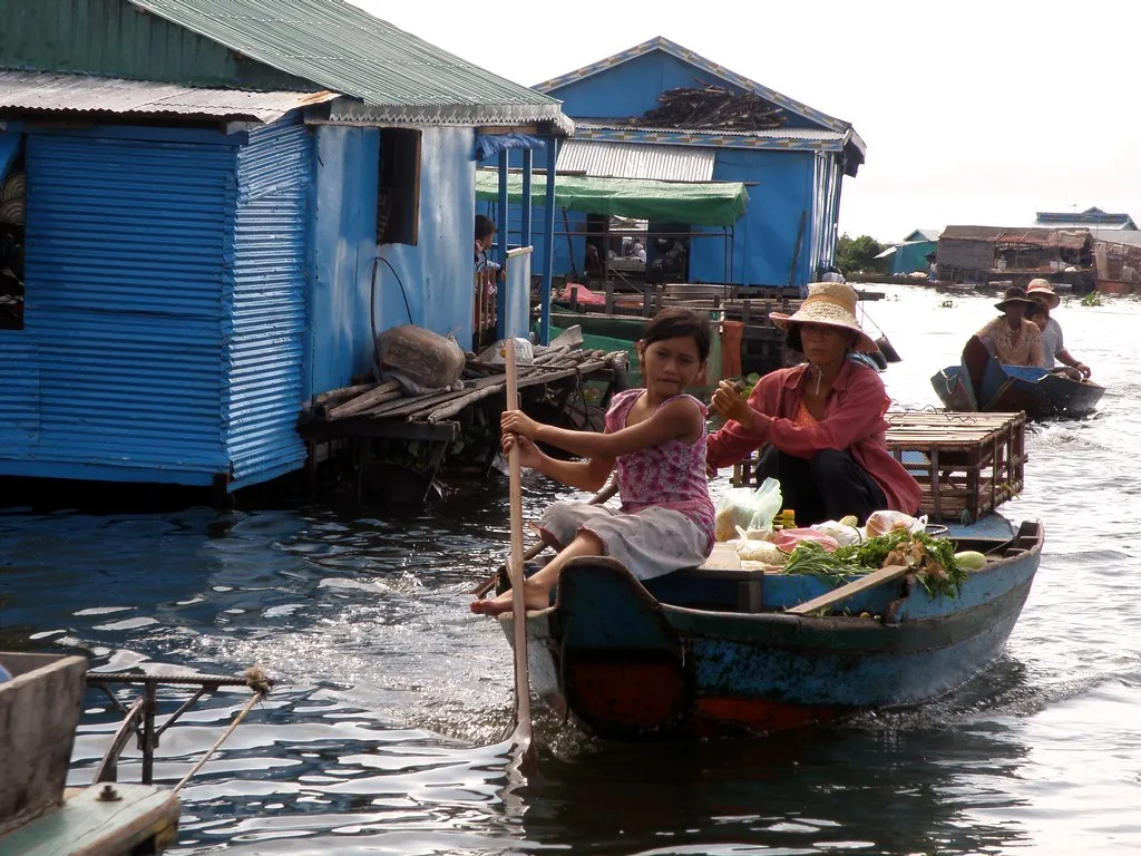 Villagers paddling a small wooden boat past stilt houses on a Cambodian river